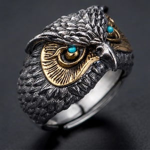 May include: A detailed ring shaped like an owl head, featuring a dark oxidised silver finish on the textured feathers and a contrasting gold-tone finish around the eyes. The owl has two small, bright turquoise-coloured circular stone inlays.