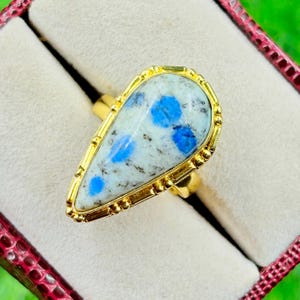 Op de afbeelding: Een goudkleurige ring met een druppelvormige steen. De steen heeft een lichtblauwe en witte achtergrond met blauwe vlekken. De ring is gezet in een goudkleurige bezel met een decoratieve rand. De ring wordt gepresenteerd in een rode sieradendoos.