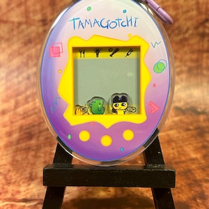 Op de afbeelding: Een vintage Tamagotchi digitaal huisdier speelgoed, met een paars en blauw ovaal ontwerp met de tekst "Tamagotchi". Het scherm toont twee geanimeerde karakters. Het heeft een paarse sleutelhanger en staat op een kleine zwarte standaard.