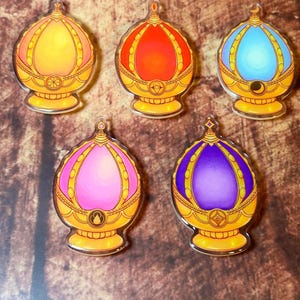 Pode incluir: Cinco pingentes de acrílico coloridos em forma de joia. Cada pingente apresenta um gradiente de cores, incluindo laranja, vermelho, azul, rosa e roxo, envolto em uma moldura dourada e ornamentada. Os pingentes têm um pequeno laço na parte superior para fixação.