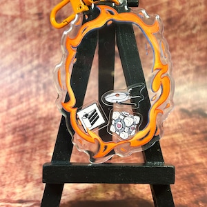 May include: A clear acrylic keychain with an orange and blue flame design. The keychain features three small images: a spaceship, a cube with hearts, and a document. The keychain is attached to an orange clasp.