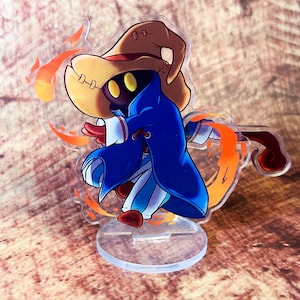 May include: A clear acrylic figurine of a cartoon character in a blue robe and a brown hat. The character has yellow eyes and is surrounded by orange flames. The figurine is on a clear circular base.
