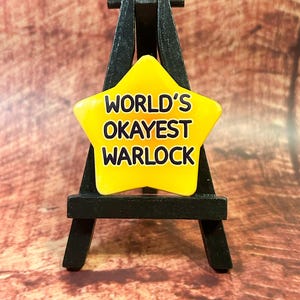 May include: A yellow star-shaped badge with the text "WORLD'S OKAYEST WARLOCK" in black lettering. The badge is displayed on a small black easel. The background is a blurred, reddish-brown wooden surface.