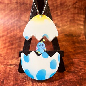 May include: A novelty keychain depicting a cartoon character hatching from an egg. The eggshell is white with blue spots, and the character is blue with large eyes. The keychain hangs from a small black display stand.
