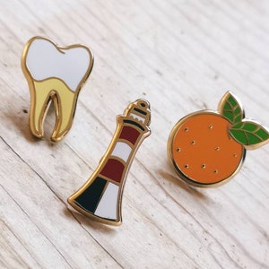 Può includere: Tre spille in smalto raffiguranti un dente, un faro e un'arancia. Il dente è giallo e bianco, il faro è rosso, bianco e nero, e l'arancia è arancione e verde.