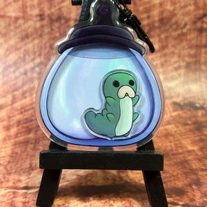 Op de afbeelding: Een heldere acryl bedel met een groene cartoonrups in een blauw getinte, met water gevulde container. De bedel heeft een zwarte sluiting en is gemonteerd op een kleine zwarte ezel. De rups heeft een witte buik en een lichtgroen lichaam.