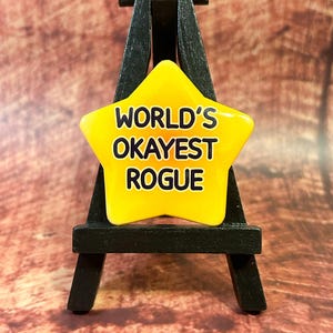 May include: A yellow star-shaped pin with the text "WORLD'S OKAYEST ROGUE" in black lettering. The pin is displayed on a small black easel against a brown wooden background. The pin is a fun, novelty item.