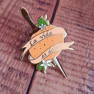 Puede incluir: Pin de esmalte dorado con una naranja estilizada atravesada por una daga. La naranja tiene el texto "La vida es dolor" escrito en ella. La daga tiene una flor en el mango.