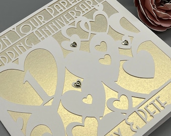 May include: A white card with a gold background and a cut-out design of hearts. The card says "On Your Paper Wedding Anniversary" and "Kelly & Pete".