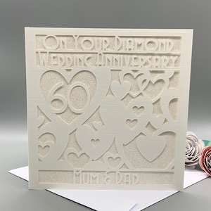 May include: White card with a cut-out design of hearts and the number 60. The text "On Your Diamond Wedding Anniversary" and "Mum & Dad" are also cut out.