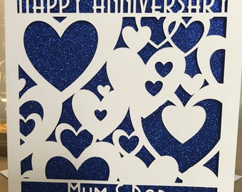 May include: A white card with a blue glitter background and a cut-out design of hearts. The card says "Happy Anniversary Mum & Dad" in white letters.