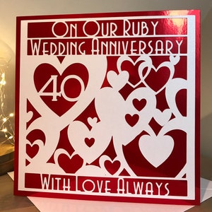 May include: Red card with a white cut-out design. The card says "On Our Ruby Wedding Anniversary" with the number 40 in a heart shape. The card also says "With Love Always".