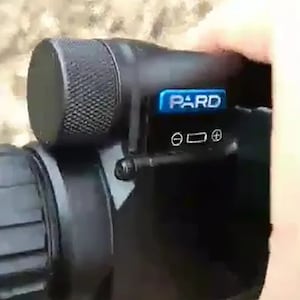 May include: A black night vision scope with a blue "PARD" logo and a black knob on the top. The scope has a black lens and a black body.