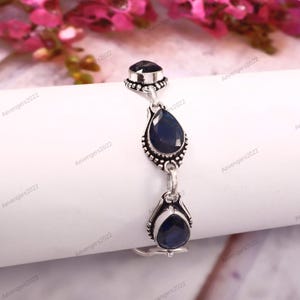 May include: A silver bracelet with three dark blue gemstones. The bracelet has an ornate design with a square stone at the top and two teardrop-shaped stones. The gemstones are set in a detailed silver setting.