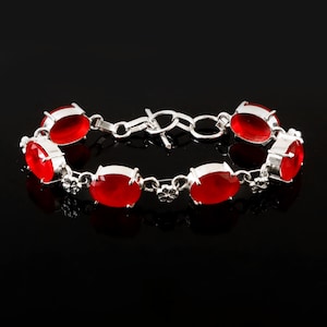 Op de afbeelding: Een zilveren armband met ovale, helderrode edelstenen. De edelstenen zijn gezet in zilver en verbonden door een delicate ketting met een toggle sluiting. Kleine bloemaccenten zijn zichtbaar tussen de edelstenen. De armband wordt getoond tegen een zwarte achtergrond.