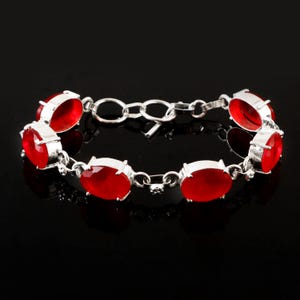 Può includere: Un bracciale in argento con pietre preziose rosse brillanti di forma ovale. Ogni pietra è incastonata in una lunetta d'argento e collegata da una sottile catena d'argento. Mostrato su sfondo nero.