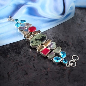 Könnte beinhalten: Ein silbernes Armband mit einer Vielzahl von bunten Edelsteinen, darunter Grün, Blau, Rot und Braun. Das Armband hat einen Verschluss.