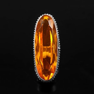 May include: A large, oval-shaped, orange gemstone ring with a silver setting. The ring has a detailed, textured design around the gemstone.