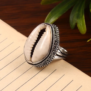 Puede incluir: Un anillo de plata con una concha de cauri blanca en el centro. El anillo tiene una banda de plata adornada con un diseño decorativo.