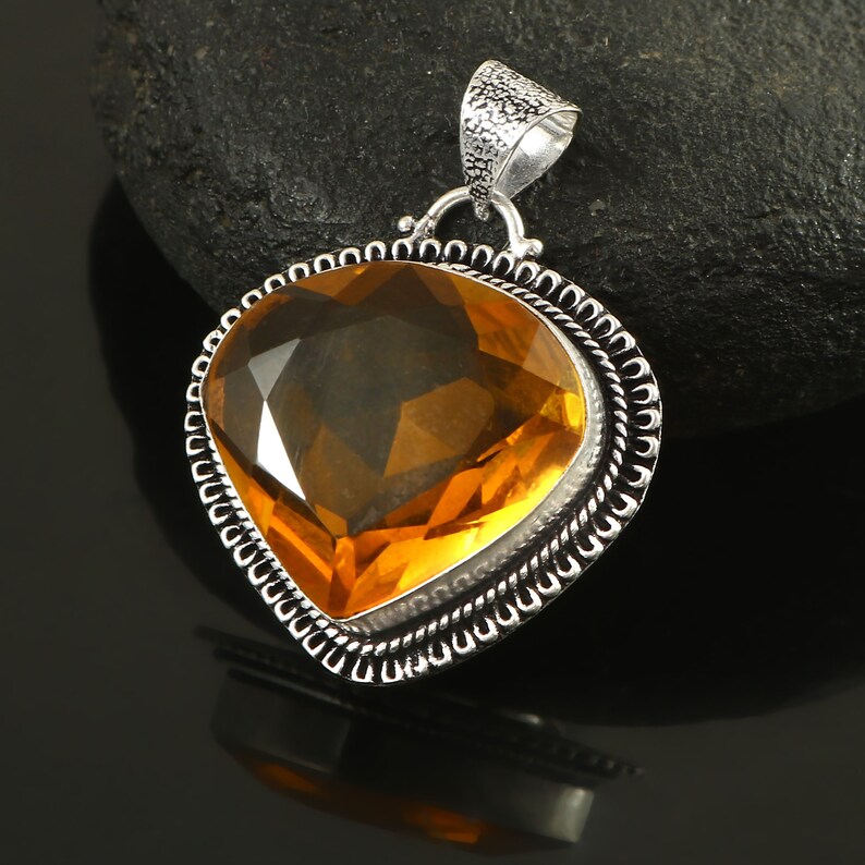 K&ouml;nnte beinhalten: Ein herzf&ouml;rmiger Anh&auml;nger mit einem gro&szlig;en, facettierten, bernsteinfarbenen Edelstein. Der Edelstein ist in einem silberfarbenen, verzierten Rahmen mit einer strukturierten &Ouml;se gefasst. Der Anh&auml;nger wird vor einem dunklen Hintergrund pr&auml;sentiert.