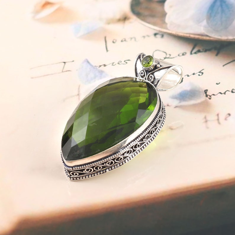 May include: A silver pendant with a large, faceted, green gemstone set in a filigree design. The pendant is on a white surface.