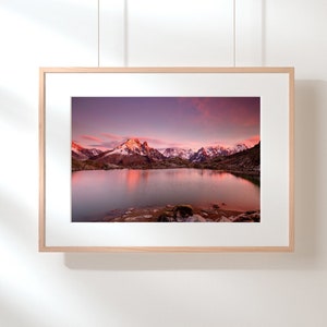 May include: A framed photograph of a mountain lake reflecting the pink and purple hues of a sunset sky. The mountains in the background are snow-capped and the water is still and calm.