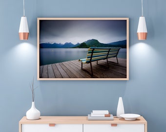 May include: A framed photograph of a wooden bench on a jetty overlooking a lake with mountains in the background. The sky is cloudy and the water is calm.