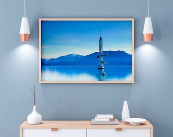 May include: A framed photograph of a blue lake with a tall metal structure in the centre. The lake is surrounded by mountains in the background. The photo is hanging on a light blue wall with two pendant lights on either side.
