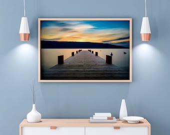 May include: A framed photograph depicting a wooden jetty stretching out into a tranquil lake at sunset. The sky is a vibrant orange and pink, with clouds mirrored in the water. The jetty is constructed from weathered wooden planks and leads towards a small island in the distance.