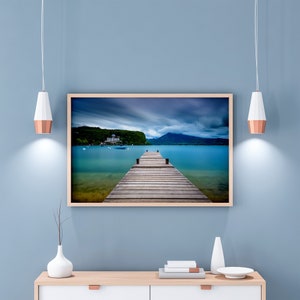 May include: A framed landscape photograph shows a wooden pier stretching into a turquoise lake, with mountains in the background under a cloudy sky. The image is displayed on a light cabinet against a blue wall, with two pendant lights.