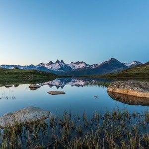 May include: A still mountain lake reflecting the snow-capped peaks of a mountain range. The lake is surrounded by green grass and rocks.