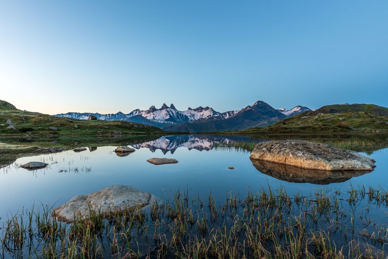 May include: A still mountain lake reflecting the snow-capped peaks of a mountain range. The lake is surrounded by green grass and rocks.