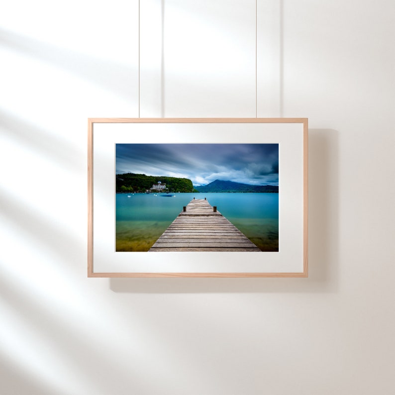 May include: A framed landscape photograph showing a wooden pier stretching into a calm turquoise lake. The sky is overcast with dramatic clouds. A building and mountains are visible in the background. The frame is light-coloured wood.