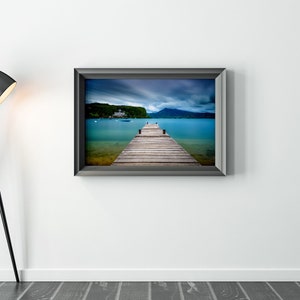 May include: A framed photograph showing a wooden pier stretching into a turquoise lake, with a building and mountains in the distance under a cloudy sky. A black floor lamp is on the left.