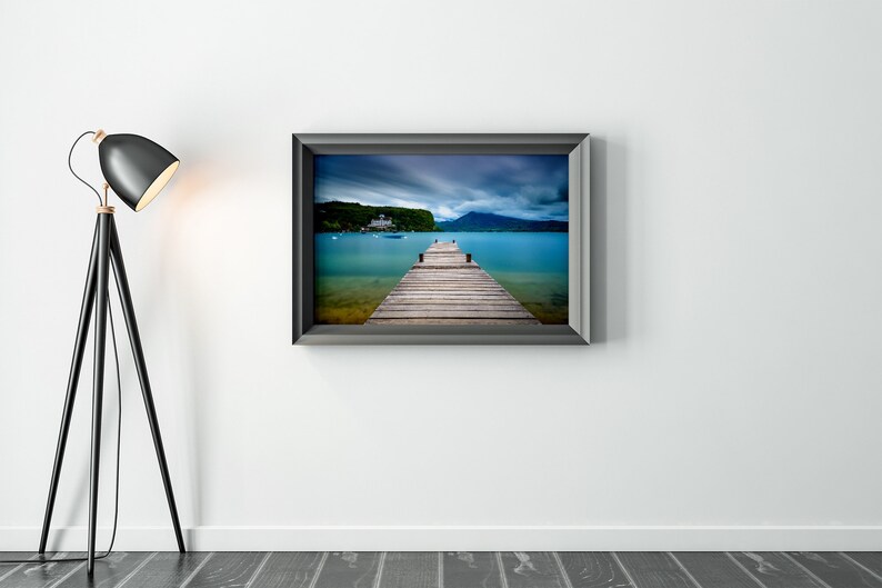 May include: A framed photograph showing a wooden pier stretching into a turquoise lake, with a building and mountains in the distance under a cloudy sky. A black floor lamp is on the left.
