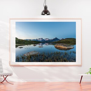 May include: A framed photograph of a mountain range reflected in a still lake. The mountains are snow-capped and the lake is surrounded by green grass and rocks.