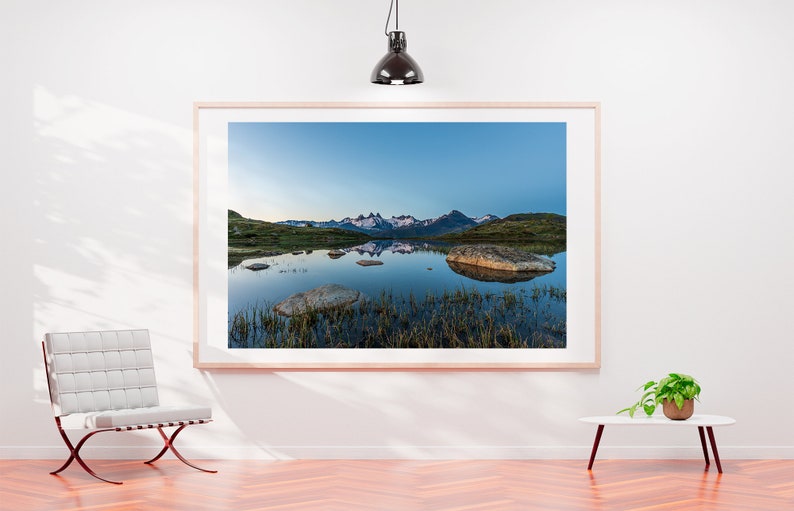 May include: A framed photograph of a mountain range reflected in a still lake. The mountains are snow-capped and the lake is surrounded by green grass and rocks.