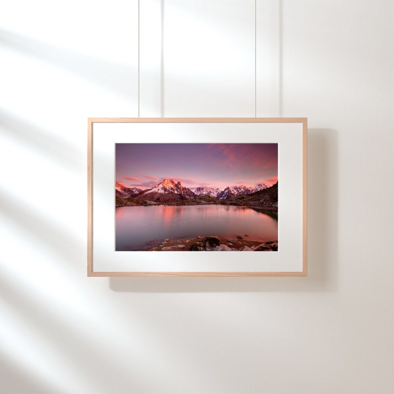 May include: A framed photograph of a mountain lake reflecting the pink and purple hues of a sunset sky. The mountains in the background are snow-capped and the water is still and calm.