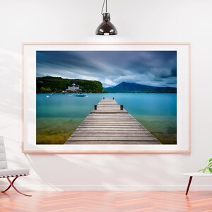 May include: A framed photograph depicting a wooden pier stretching into a turquoise lake, with mountains and a cloudy sky. The artwork hangs on a white wall, complemented by a modern chair and a small table with a plant.
