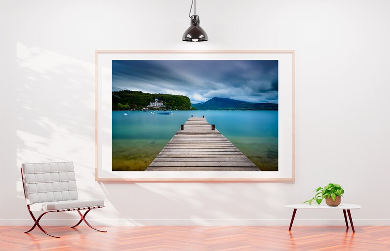 May include: A framed photograph depicting a wooden pier stretching into a turquoise lake, with mountains and a cloudy sky. The artwork hangs on a white wall, complemented by a modern chair and a small table with a plant.