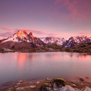 May include: A scenic view of a mountain lake with snow-capped peaks reflected in the still water. The sky is a vibrant pink and purple, creating a beautiful sunset over the mountains.