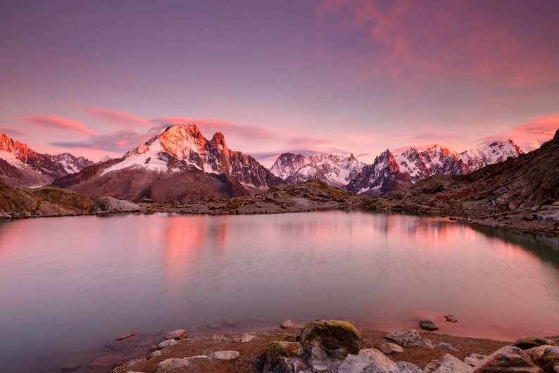 May include: A scenic view of a mountain lake with snow-capped peaks reflected in the still water. The sky is a vibrant pink and purple, creating a beautiful sunset over the mountains.
