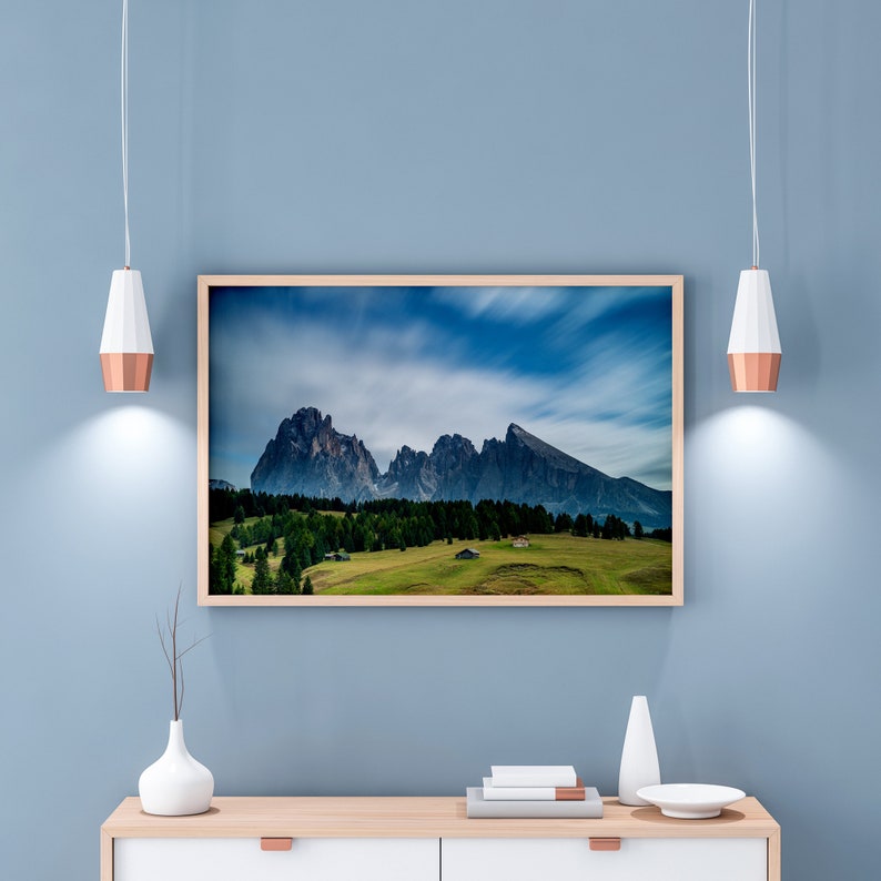 May include: A framed landscape print featuring a mountain range under a cloudy sky. The print is in a light wood frame and hangs on a blue wall above a wooden cabinet. The scene includes green trees and a grassy field.