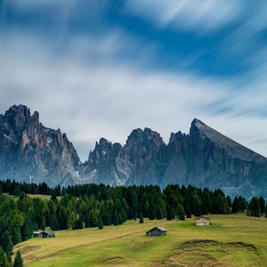 May include: A scenic landscape featuring a range of jagged, gray mountains under a cloudy blue sky. Lush green trees and grassy fields fill the foreground, with small buildings scattered throughout the valley. The image captures a serene, natural environment.