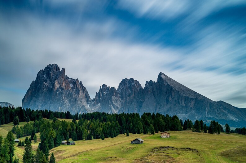 May include: A scenic landscape featuring a range of jagged, gray mountains under a cloudy blue sky. Lush green trees and grassy fields fill the foreground, with small buildings scattered throughout the valley. The image captures a serene, natural environment.
