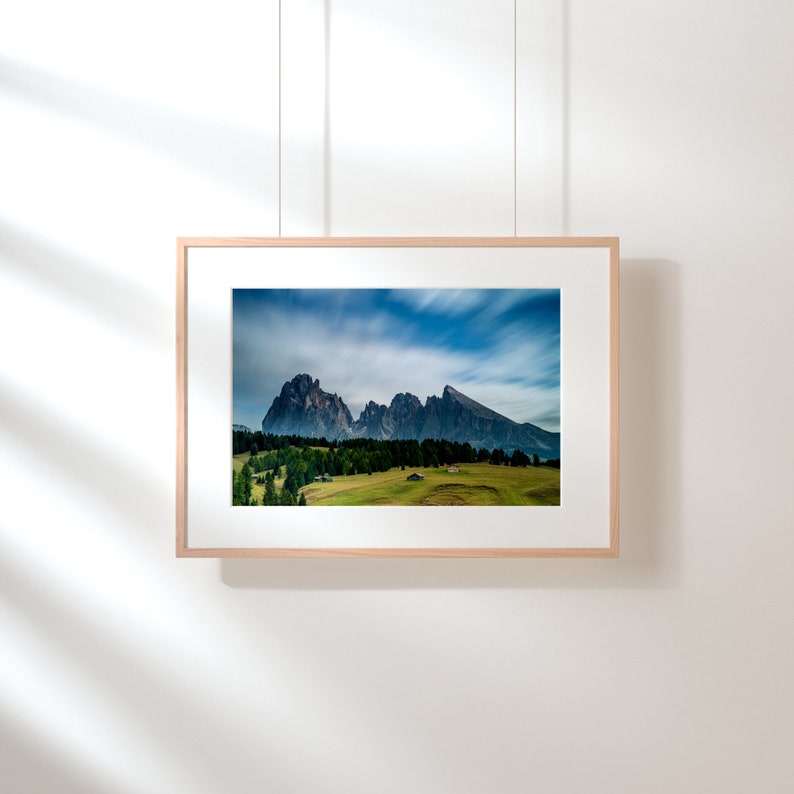 May include: A framed landscape photograph featuring a mountain range under a dynamic, cloudy sky. The image showcases a green meadow with trees and small buildings. The frame is made of light-colored wood, and the photograph is suspended by thin cords.