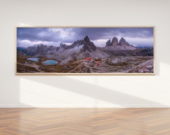May include: A panoramic photograph of a mountain range with a small building and two lakes in the foreground. The sky is cloudy and the mountains are rocky and snow-capped.