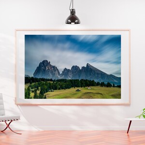 May include: A framed landscape photograph of a mountain range under a cloudy blue sky. The image features green fields, trees, and small buildings at the base of the mountains. The frame is light-colored, and the photograph is displayed on a white wall.