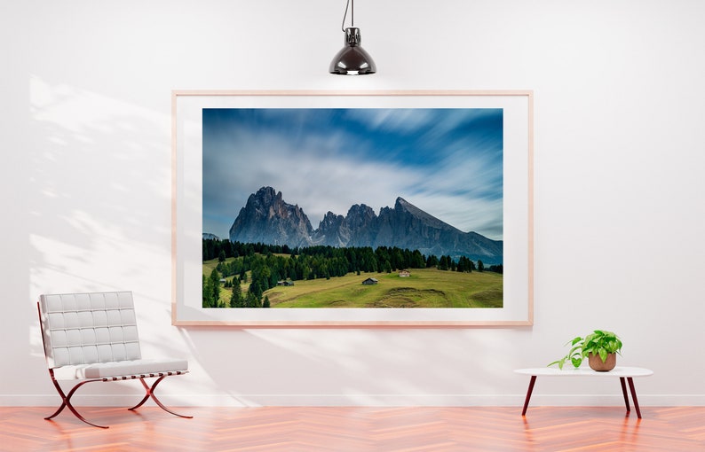 May include: A framed landscape photograph of a mountain range under a cloudy blue sky. The image features green fields, trees, and small buildings at the base of the mountains. The frame is light-colored, and the photograph is displayed on a white wall.