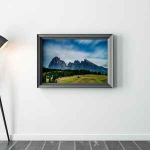 May include: A framed landscape photograph of a mountain range with a green valley and a blue sky. A black floor lamp with a black lampshade is to the left of the framed photograph.